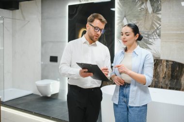 Satıcı elinde bir pano ile modern seramik fayans ve tesisat dükkanındaki müşterilere banyo mobilya seçeneklerini gösteriyor.