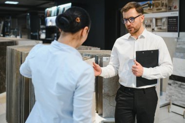 Kadın müşteri, iyi aydınlatılmış seramik fayans ve tesisat mağazasında çeşitli fayans örneklerini sergileyen erkek danışman ile seçenekleri tartışıyor.