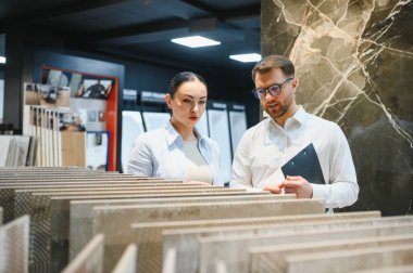 Bir hırdavatçıda kadın müşteriye seramik fayans örneklerini gösterirken elinde bir pano tutan satıcı, ev geliştirme seçeneklerini tartışıyor.
