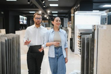 Satıcının elinde bir pano var. Parlak, modern bir hırdavatçıda gülümseyen bir müşteriye çeşitli seramik fayanslar sunuyor.