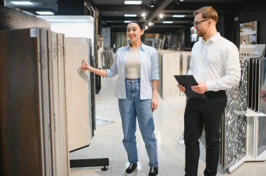 Dükkan asistanı elinde bir pano tutuyor, bir kadına çeşitli stil ve renklerle dolu bir perakende mağazasında seramik fayanslar seçmesini tavsiye ediyor.