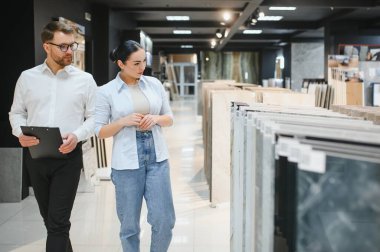 Nalbur dükkanındaki kadın müşteriye seramik fayanslar gösteren bir pano tutan satıcı yenileme projesi için fayansları seçiyor.