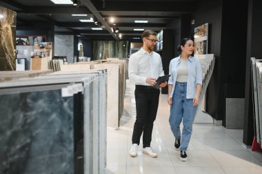 Satıcının elinde bir pano var. Hırdavatçıdan geçen bir kadına çeşitli seramik fayanslar gösteriyor.