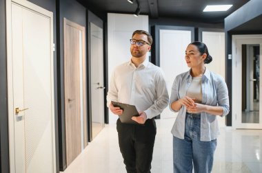 Satıcının elinde pano var. Kapıdaki ve pencerelerdeki kadın müşterilere farklı kapılar gösteriyor.