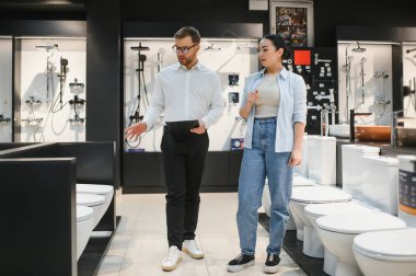 Satış elemanı, su tesisatı mağazasında çalışan bir kadına farklı tuvalet modelleri gösteriyor. Doğru olanı seçmesine yardım ediyor.
