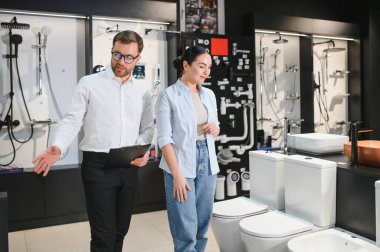 Satıcı kadın müşterilere farklı banyo armatürleri gösteriyor. Tesisat mağazasında pano tutuyor, seramik klozet ya da lavabo seçiyor.