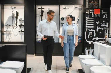 Kadın müşteri ve erkek satıcı modern bir mağazada seramik fayanslar ve tesisat demirbaşlarını tartışıyor, çeşitli banyo çözümleri sergiliyorlar.