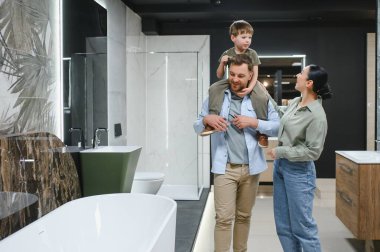 Mutlu bir aile, seramik fayanslara ve hijyenik malzemelere bakarken oğullarını omuzlarında taşıyarak bir tesisat dükkanında yeni banyo eşyalar seçiyor.