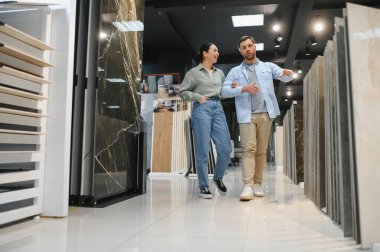 Fayans ve tesisat dükkanında yürüyen bir çift ev yenileme seçeneklerini tartışıyor. Çeşitli seramik fayanslar ve tesisat malzemeleri sergileniyor