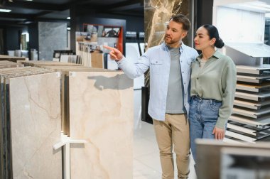 Bir ev geliştirme mağazasında seramik fayans örneğine işaret eden çift, yenileme için malzeme tartışıyor ve seçiyor
