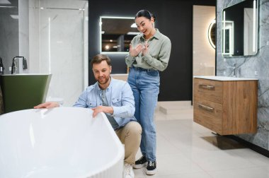 Tesisatçı dükkanında seramik küveti seçen mutlu çift, gülümseyen kadın ve erkek galeride banyo mobilyalarını seçiyor.