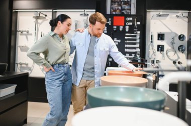 Modern bir sıhhi tesisat dükkanında çeşitli banyo armatürlerini ve seramik fayansları inceleyen çift, ev yenileme için tasarım seçeneklerini tartışıyor.