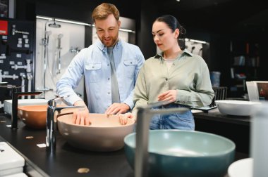 Bir çift seramik fayans ve tesisat dükkanında banyo lavabolarını inceliyor, ev yenileme veya iyileştirme seçeneklerini değerlendiriyor.