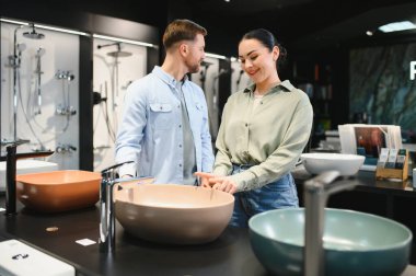 Genç çift modern tesisat mağazasındaki banyo mobilyalarını seçerken bej seramik lavaboyu işaret ediyor.