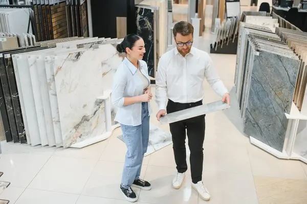 Kadın müşteri ve erkek satış uzmanı çeşitli fayans örnekleri ve tesisat malzemeleriyle çevrili modern bir mağazada seramik fayans stillerini tartışıyor.