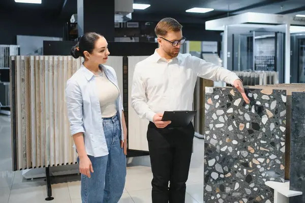 Modern seramik ve su tesisatı mağazasında erkek bir satıcıyla fayans seçeneklerini tartışan kadın müşteri çeşitli fayans tasarımları sergiliyor.