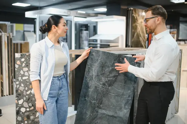 Seramik ve sıhhi tesisat dükkanında seramik fayans sunarken bir kadınla ilişki kuran tezgahtar uzman tavsiyesi ve yardımı sunuyor.