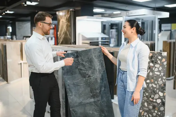 Gülümseyen tezgahtar hırdavatçıda çalışan bir kadına seramik fayans gösteriyor. Yenileme projesi için malzeme seçiyor.