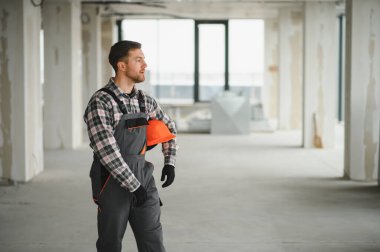 İnşaat işçisi tamamlanmamış bir binanın içinden geçiyor, elinde turuncu bir miğfer tutarak beton yapı ve inşaat çalışmalarını yürütüyor.