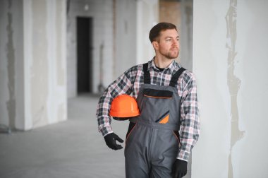 İnşaat işçisi turuncu bir kask takıyor ve inşaat halindeki bir binanın içindeki kameraya gülümsüyor.