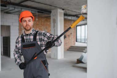 İnşaat işçisi turuncu bir kask takıyor ve inşaat halindeki bir binada duvarları boyuyor.