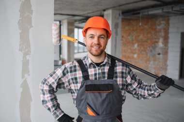 Gülümseyen müteahhit iş tulumu ve koruyucu kask giyip evin tadilatı sırasında boya silindiri tutuyor.