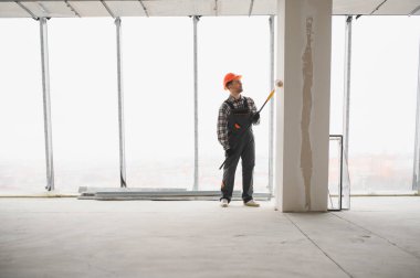 İnşaat işçisi koruyucu kask ve tulum giyiyor, uzun bir silindir kullanarak duvarı boyuyor, tadilat sırasında...