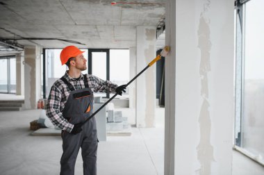 İnşaat işçisi koruyucu kask ve tulum giyiyor, uzun bir silindir kullanarak duvarı boyuyor, tadilat sırasında...