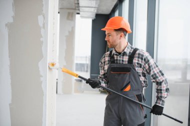 Koruyucu iş giysisi ve kask giyen inşaatçı inşaat halindeki bir binada boya silindiri kullanarak duvarlara astar takıyor.