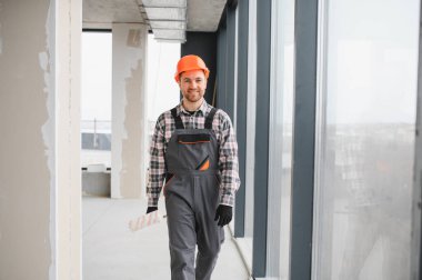 İnşaatçı kasketli ve üniformalı, inşaat alanından güvenle geçiyor, aletleri taşıyor ve gururla gülümsüyor.