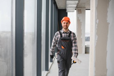 İnşaat işçisi inşaat halindeki bir binaya elinde boya silindiri, iş tulumu ve güvenlik kaskı ile giriyor.