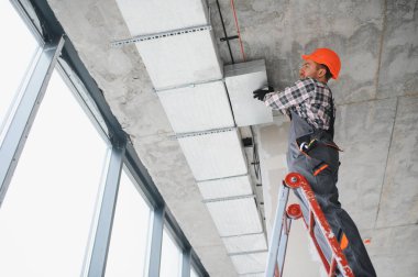 İnşaat işçisi tavana havalandırma kanalı kuruyor, inşaat halindeki bir binada merdiven kullanıyor.