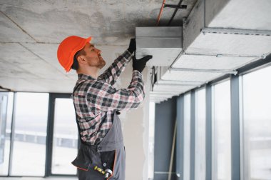 İnşaat işçisi güvenlik kaskı takıyor ve inşaat halindeki bir binaya havalandırma boruları yerleştiriyor.