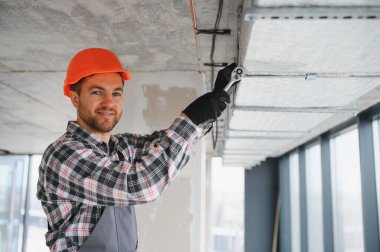 Turuncu kasklı ve işçi tulumlu bir inşaatçı inşaat halindeki bir binaya havalandırma sistemi kurarken bir vidayı sıkmak için anahtar kullanıyor.