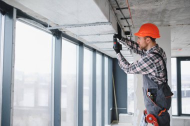 İnşaat işçisi elektrikli matkap kullanıyor, binanın tavanına havalandırma sistemi kuruyor.