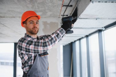 Güvenlik kaskı takan inşaat işçisi inşaat halindeki binaya havalandırma sistemi kurmak için elektrikli matkap kullanıyor.