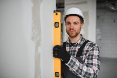 Koruyucu kask ve eldiven giyen inşaatçı, yenileme veya inşaat çalışmaları sırasında bir duvarın dikey hizalanmasını sağlamak için ruh seviyesini kullanır.