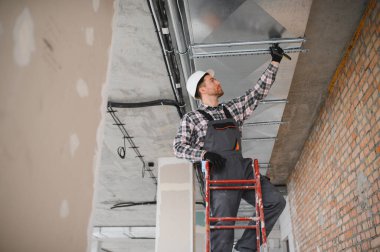 Güvenlik kaskı takan ve iş tulumu giyen inşaat işçisi merdivenin üzerinde dururken havalandırma ya da havalandırma sistemi kuruyor.