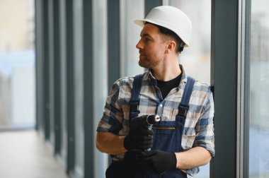 İnşaat işçisi koruyucu giysiler giyiyor ve elinde kablosuz matkap var. İnşaat alanındaki çalışmaların gidişatını kontrol ediyor.