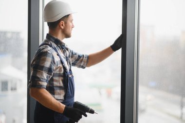 Güvenlik kaskı ve eldiven takan inşaat işçisi modern bir binaya elektrikli matkap kullanarak pencere çerçevesi yerleştiriyor.