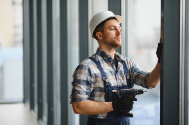 Modern binaya pencere kurarken güvenlik kaskı takan ve kablosuz matkap kullanan inşaat işçisi