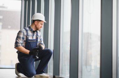 İnşaat işçisi diz çöküp modern bir binaya kablosuz tornavida kullanarak pencere kurmaya hazırlanıyor.