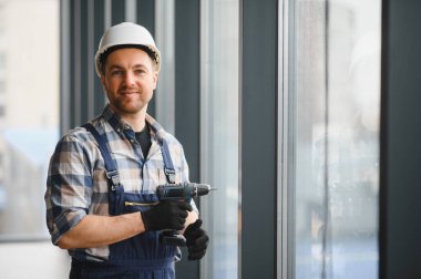 İnşaat işçisi güvenlik kaskı takıyor ve bir binaya cam yerleştirirken kablosuz matkap tutuyor.