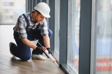 Koruyucu kask ve eldiven takan inşaat işçisi modern binanın pencere çerçevesine dolgu macunu sürüyor.