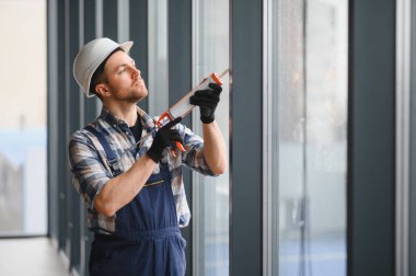 Güvenlik kaskı ve eldiven takan inşaat işçisi modern bir binanın pencere çerçevesine dolgu macunu sürüyor.