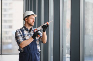 Modern bir binanın pencere kurulumu için silikon dolgu maddesi kullanan koruyucu kask ve eldiven giyen inşaat işçisi.