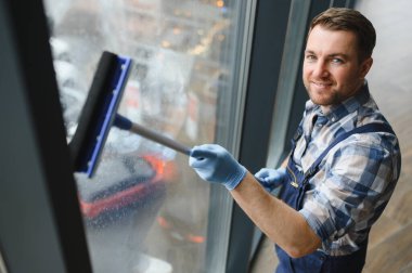 Gülümseyen temizlikçi mavi eldiven giyerek pencereleri titizlikle siliyor, ticari alanda pırıl pırıl parlıyor.