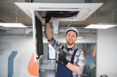 Kask takan ve elinde kablosuz matkap tutan Hvac teknisyeni, tavana monte edilmiş kaset tipi klima ünitesini tamir ediyor.