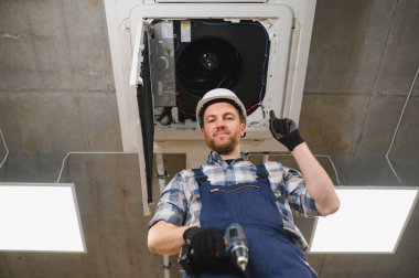 Güvenlik kaskı takan ve elinde kablosuz tornavida tutan Hvac teknisyeni, tavana monte edilmiş kaset tipi klima ünitesini tamir ediyor ve işaret veriyor.