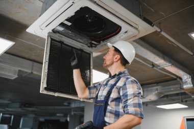 Hvac teknisyeni, koruyucu başlık ve tulum giyip bakım sırasında havalandırma ünitesinin filtresini inceliyor.